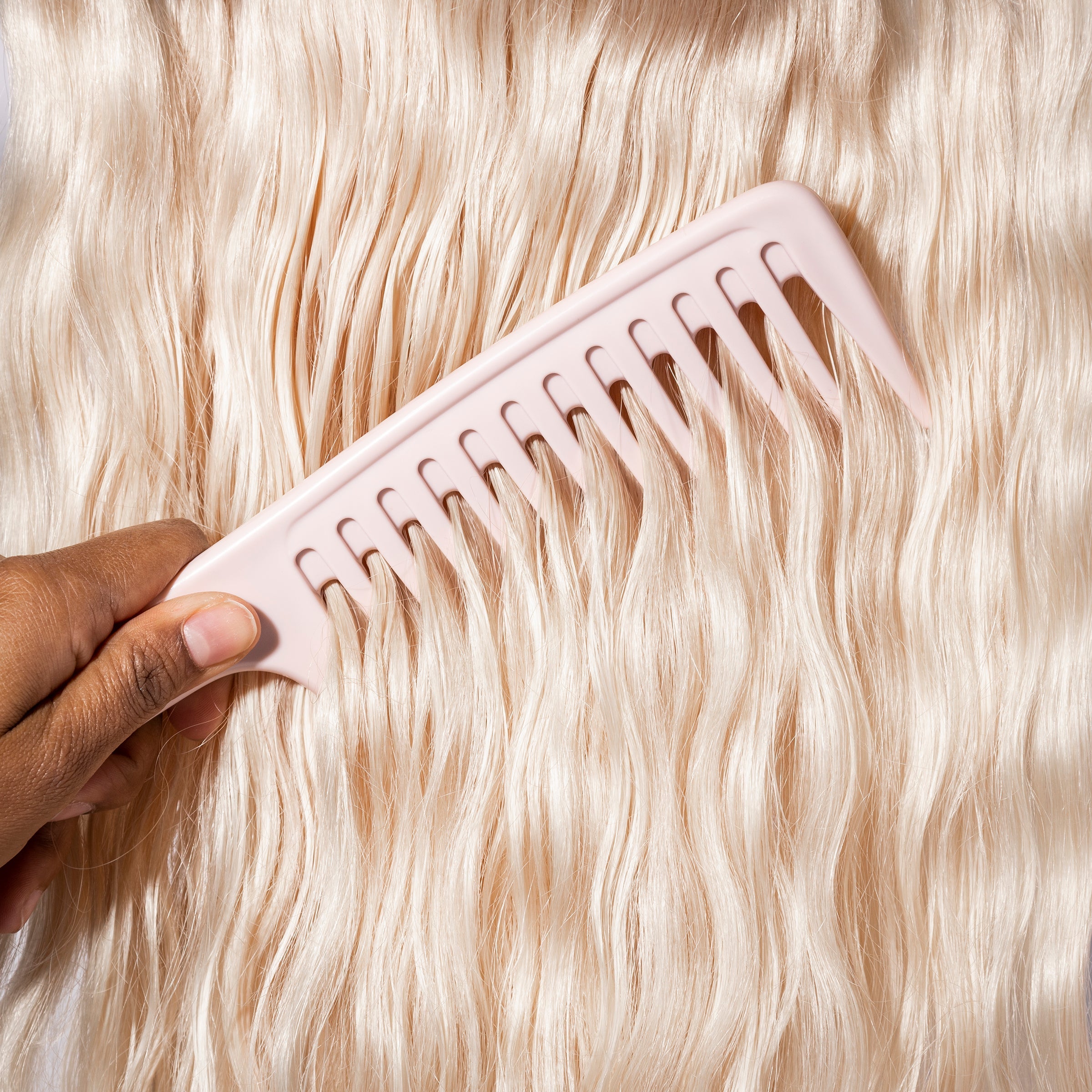 A hand gently pulls the pink detangling comb through wavy, platinum blonde hair.
