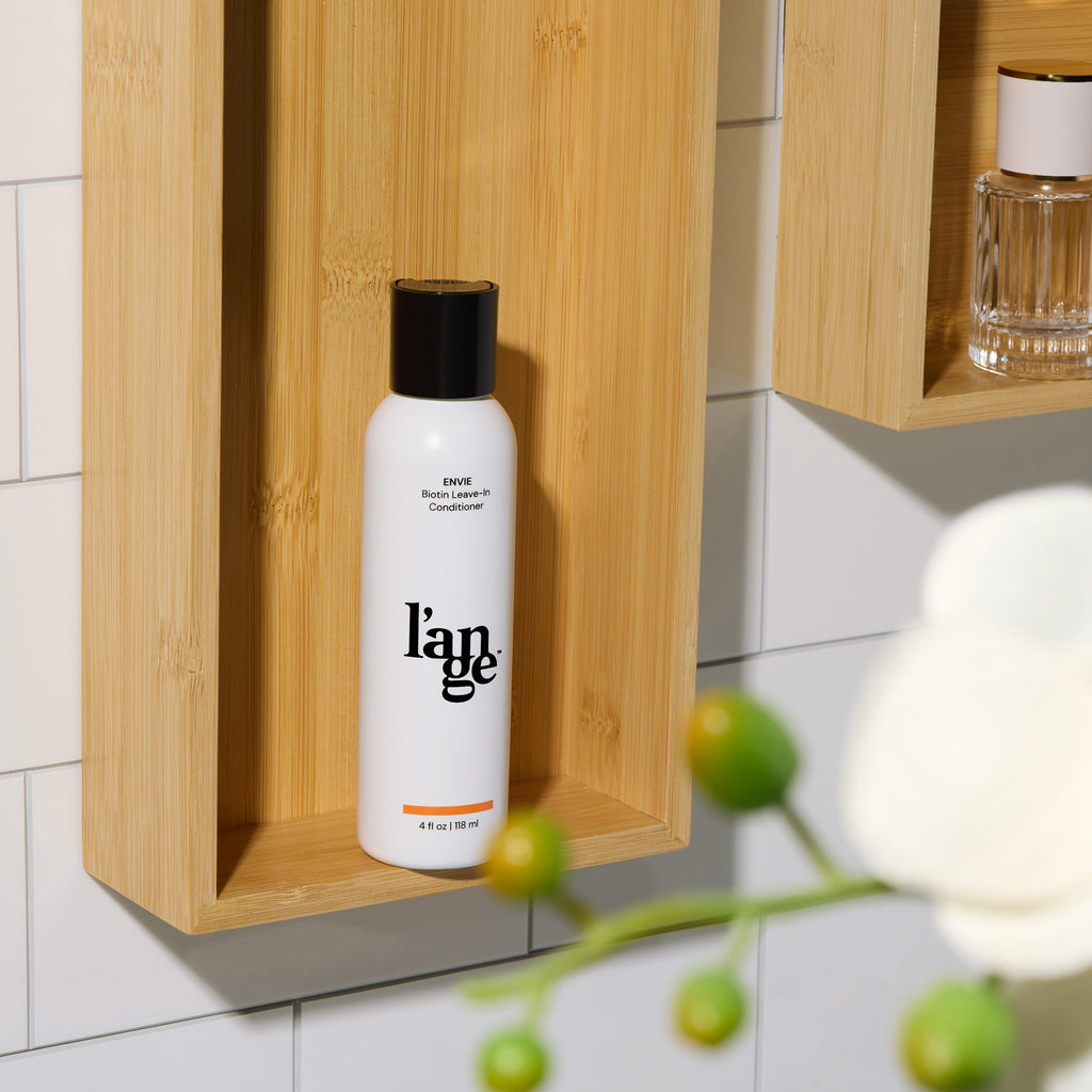 L’ange Envie Biotin Leave-In Conditioner displayed on a wooden bathroom shelf with tiled wall background.