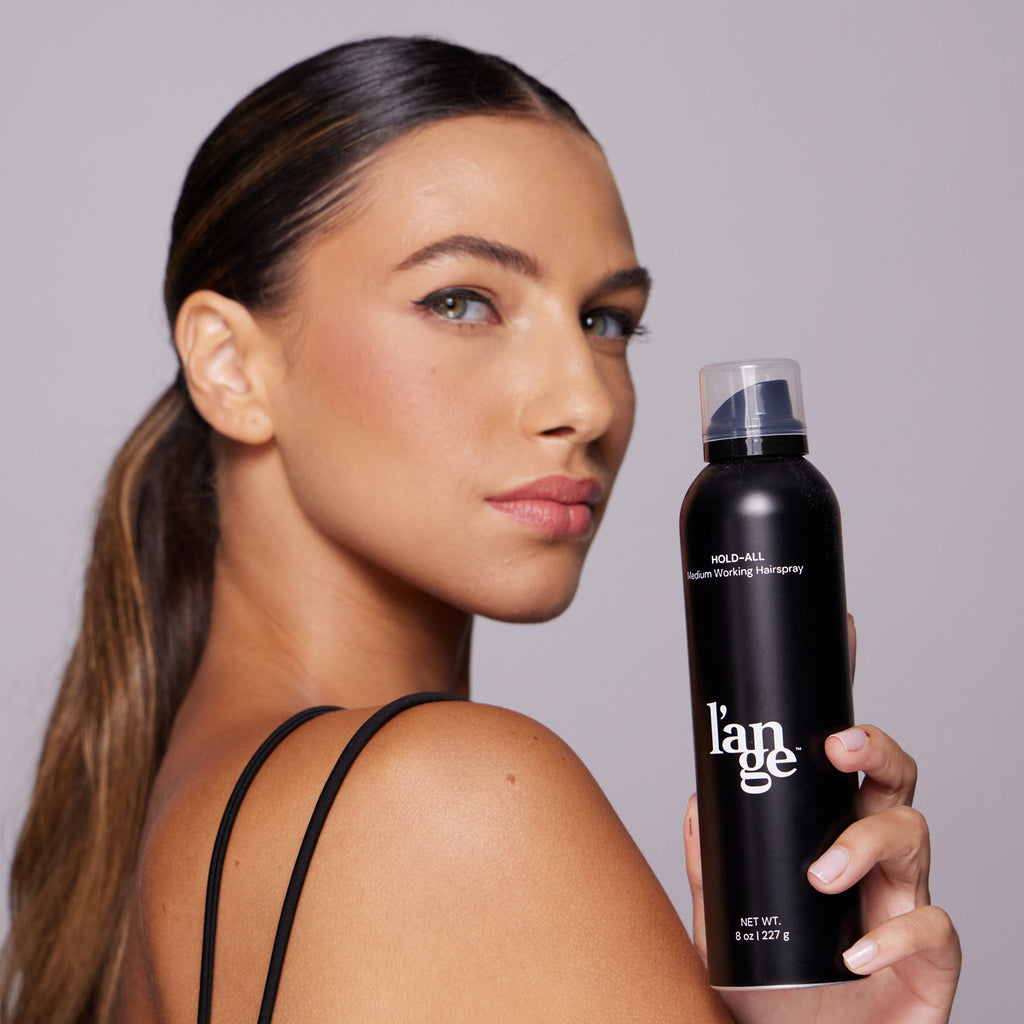 Woman with sleek ponytail holding a black can of L’ange Hold-All Medium Working Hairspray.