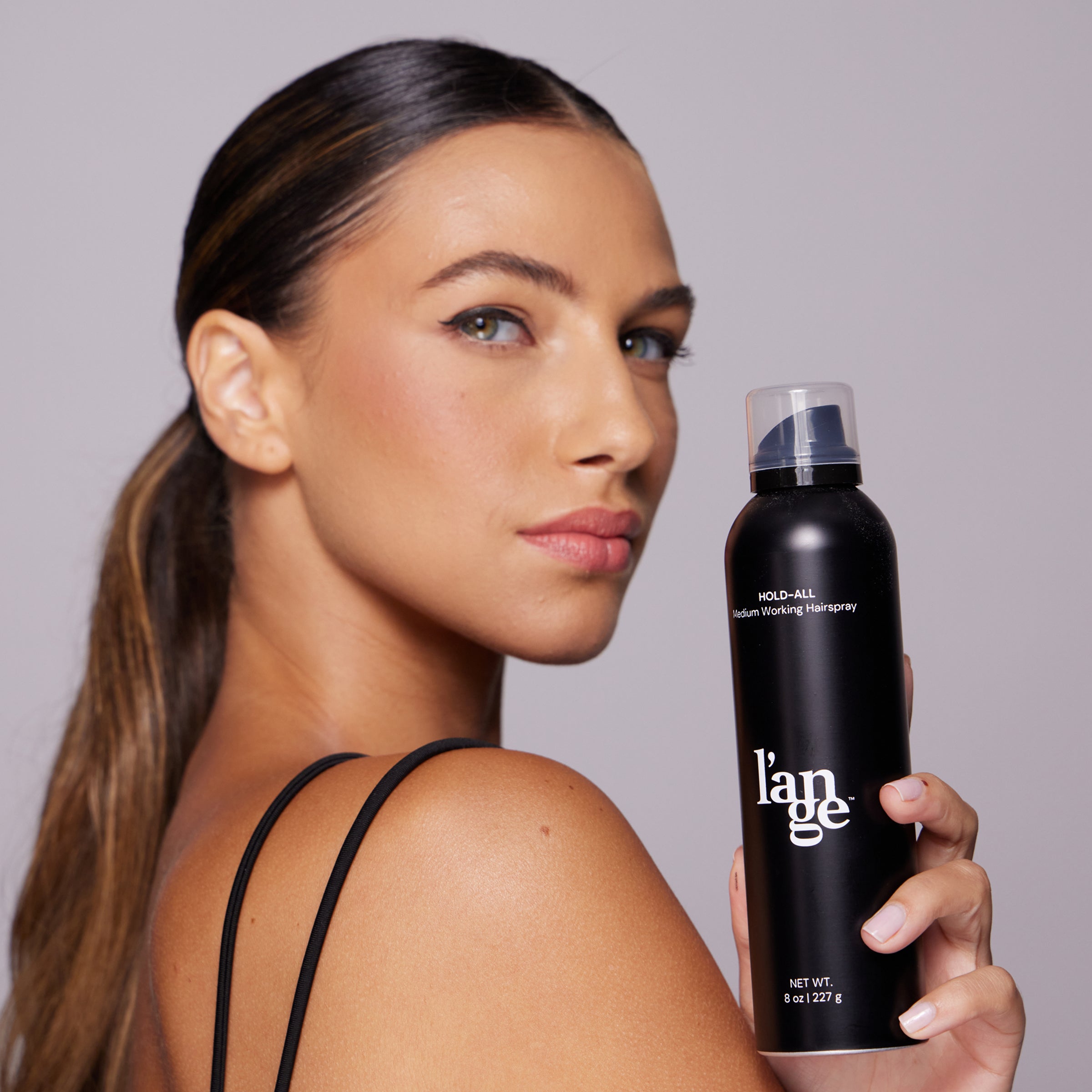 Woman with sleek ponytail holding a black can of L’ange Hold-All Medium Working Hairspray.