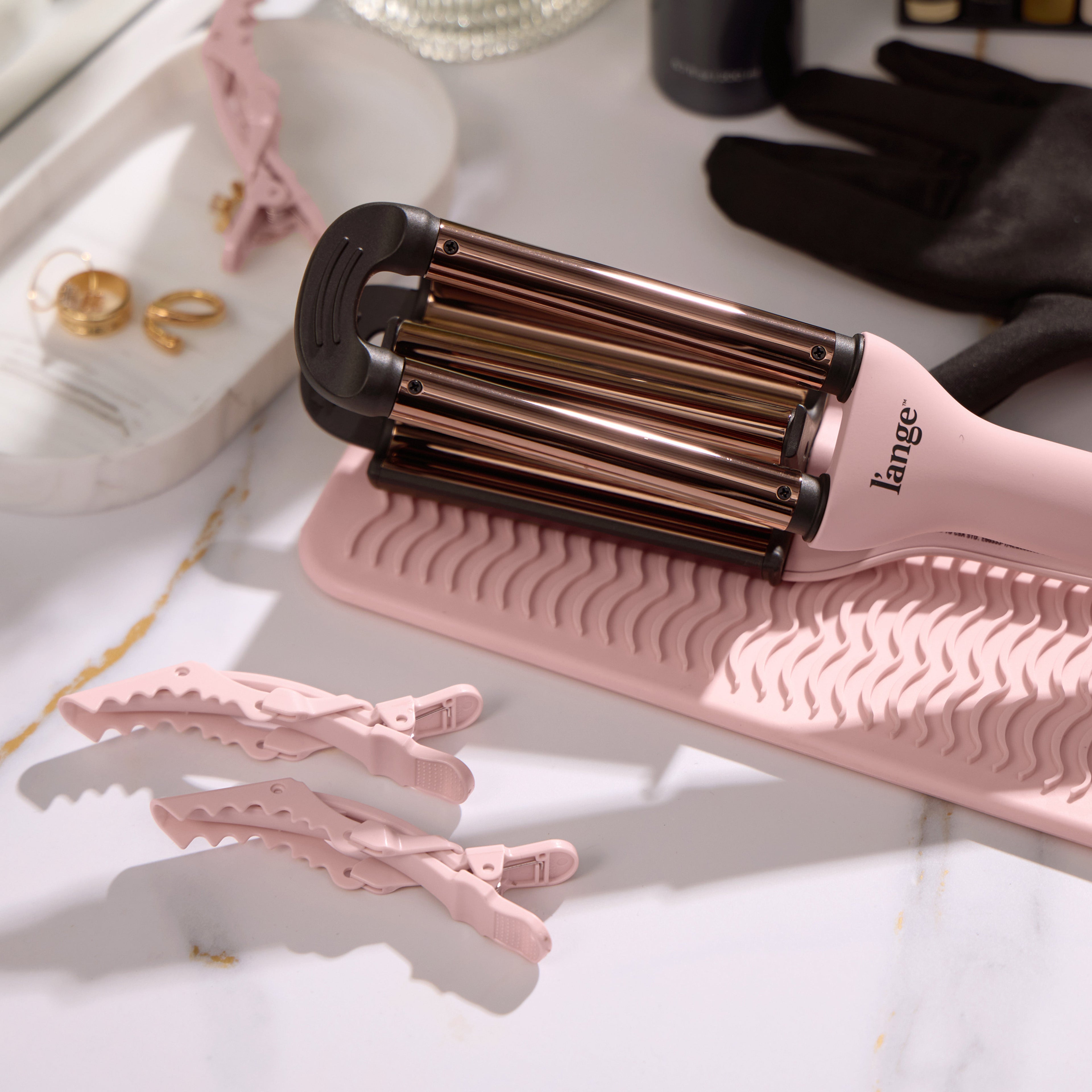 A pink and black hair curling tool rests on a wavy textured mat, accompanied by two pink hair clips and a pair of gold earrings on a nearby tray.
