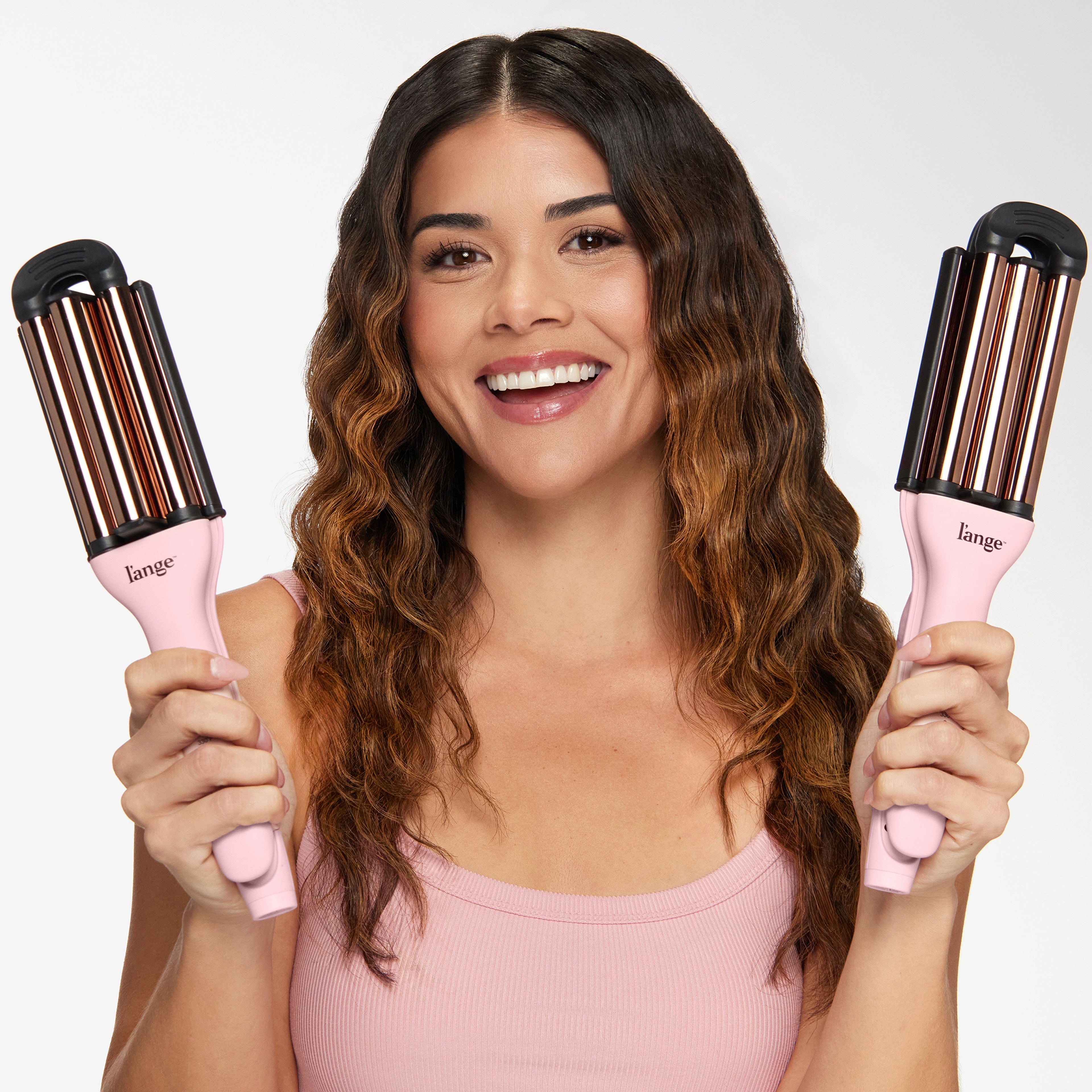 A smiling woman holds two pink, multi-barrel curling wands, showcasing her wavy hair.