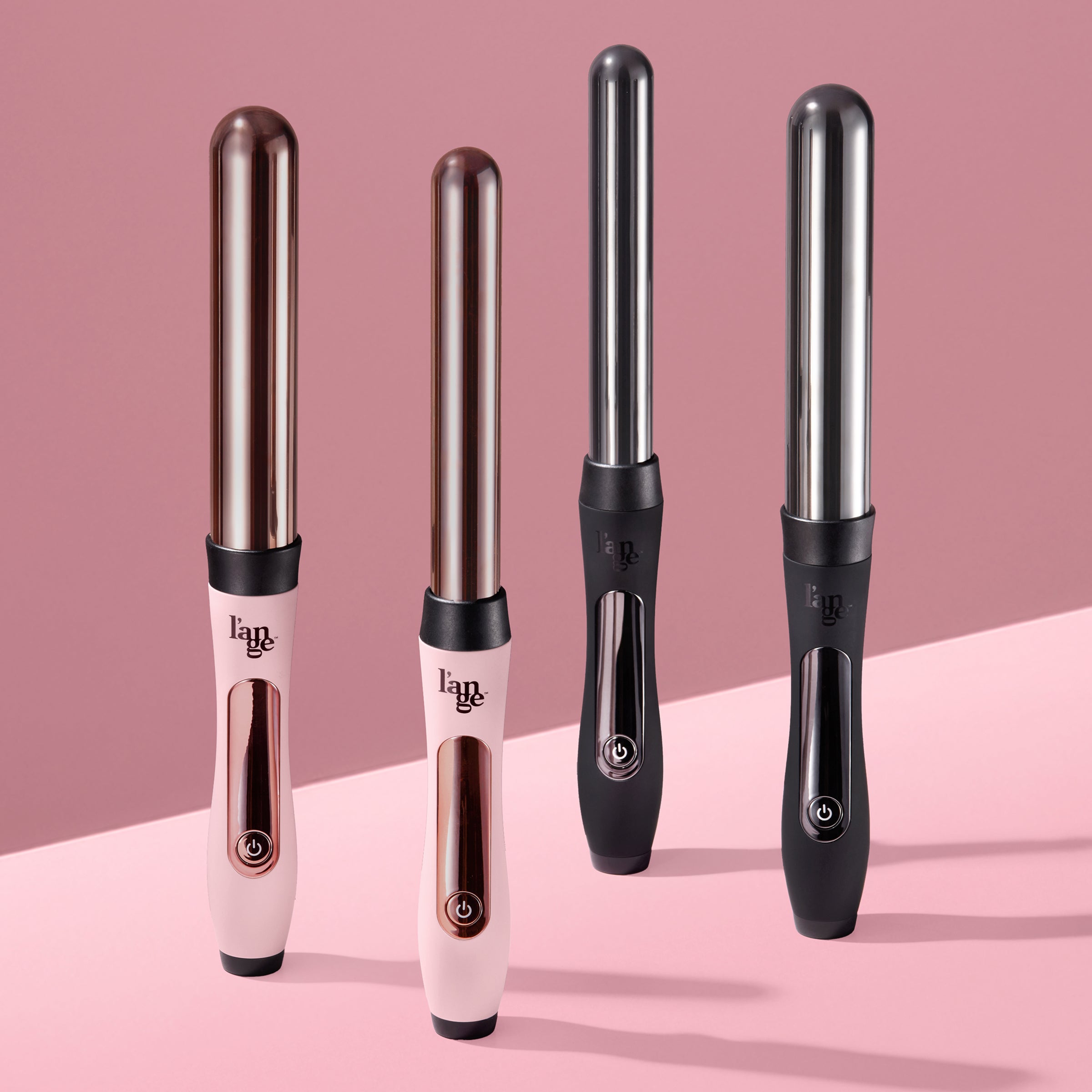 "Four L’ange curling wands standing upright on a pink surface with a pink background. Two are light pink with rose gold barrels and handles, and two are matte black with silver barrels and chrome accents.