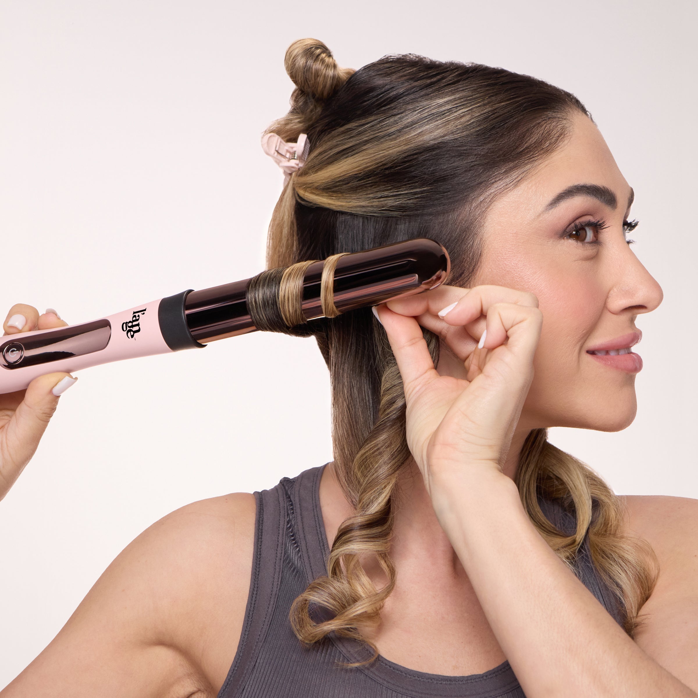 A woman with partially sectioned hair smiles while using a pink L’ange curling wand to curl a strand. Her hair is clipped up and she's wearing a gray tank top. The curling wand features a rose gold titanium barrel