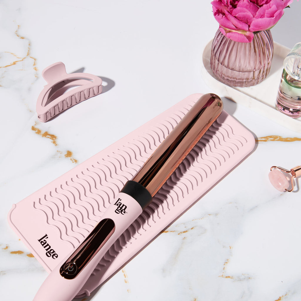 Pink and gold curling iron on a textured pink mat, accompanied by a pink hair clip and decorative pink rose in a vase, set on a marble surface.