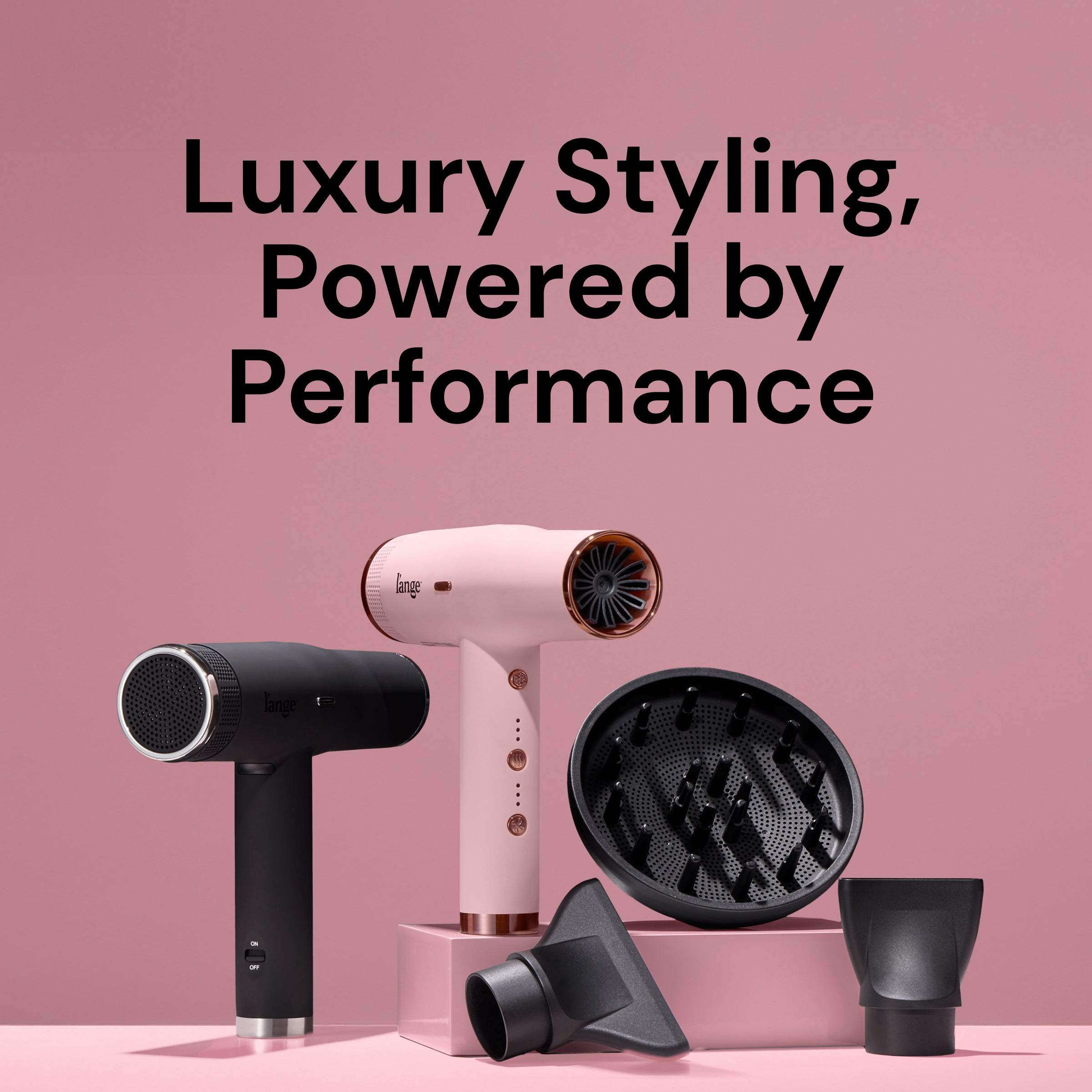 Two L’ange Le Styliste hair dryers—one in matte black and one in blush pink—displayed on a pink backdrop alongside diffuser and concentrator attachments. Large black text above reads: ‘Luxury Styling, Powered by Performance