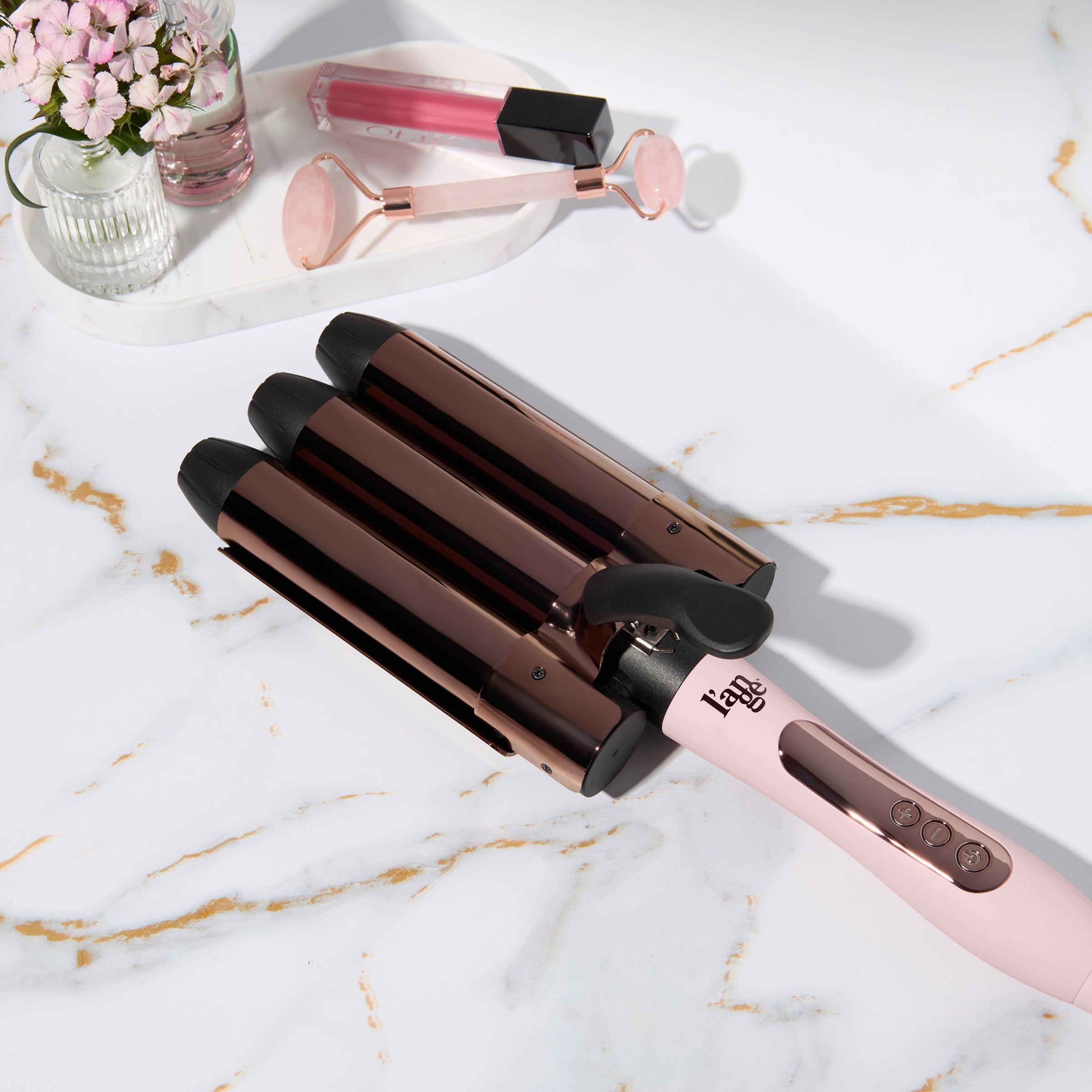 Blush triple-barrel hair waver on a marble countertop next to beauty accessories.