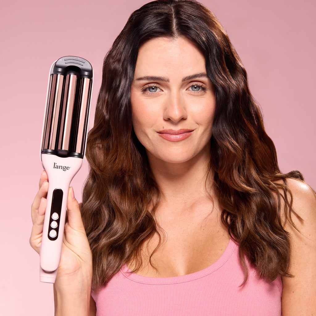 Woman with wavy brown hair holding a pink L’ange hair waver against a pink background.