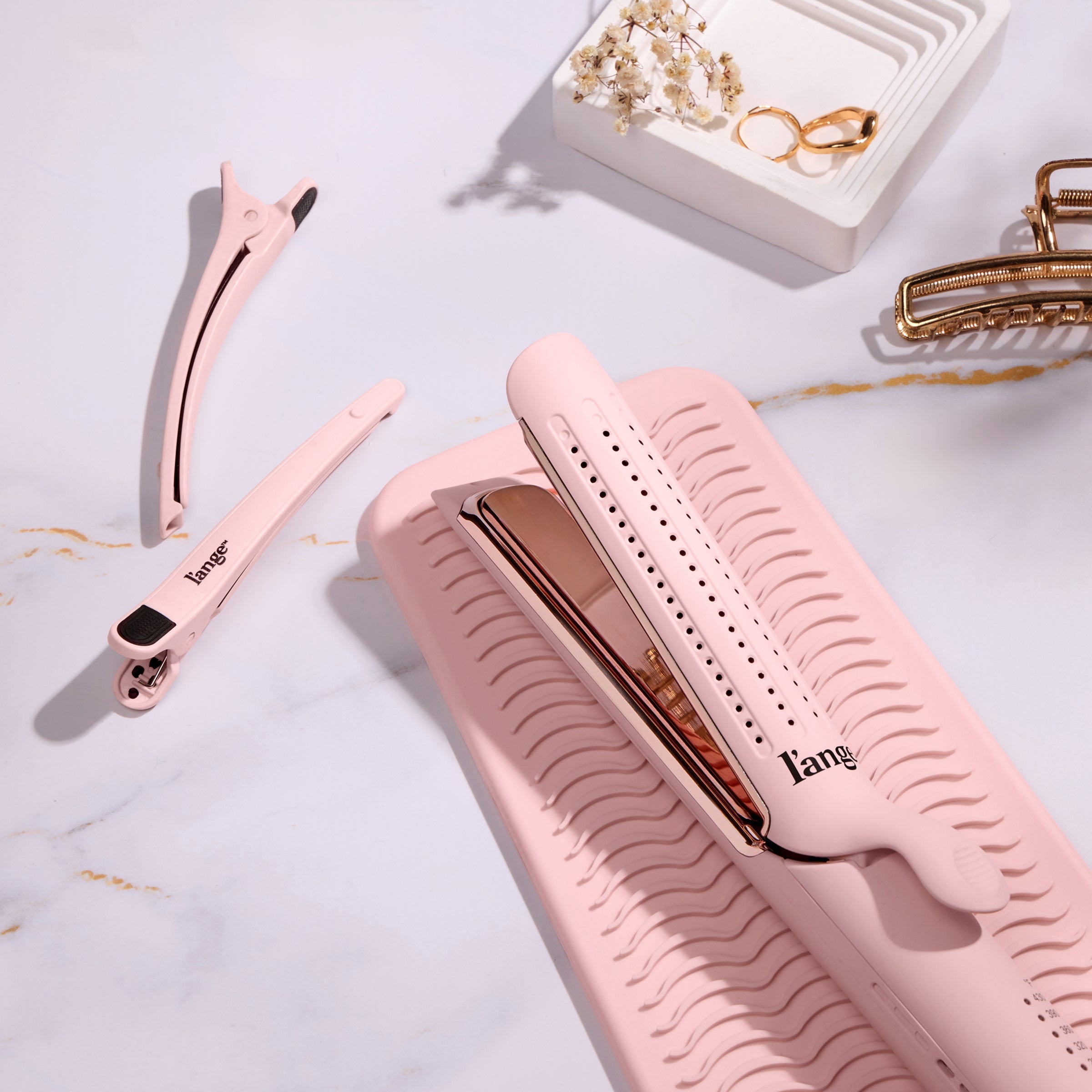 Pink hair styling set featuring a flat iron on a matching silicone mat, accompanied by a hair clip and elegant gold jewelry accents.