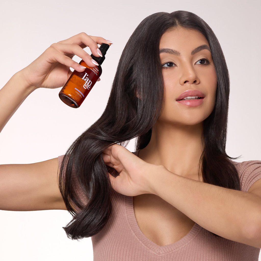 A woman with smooth, shiny hair holds a section of her hair while applying L’ange MANAGÉ Marula Oil Hydrating Mist, showcasing the product’s use for adding shine and hydration.