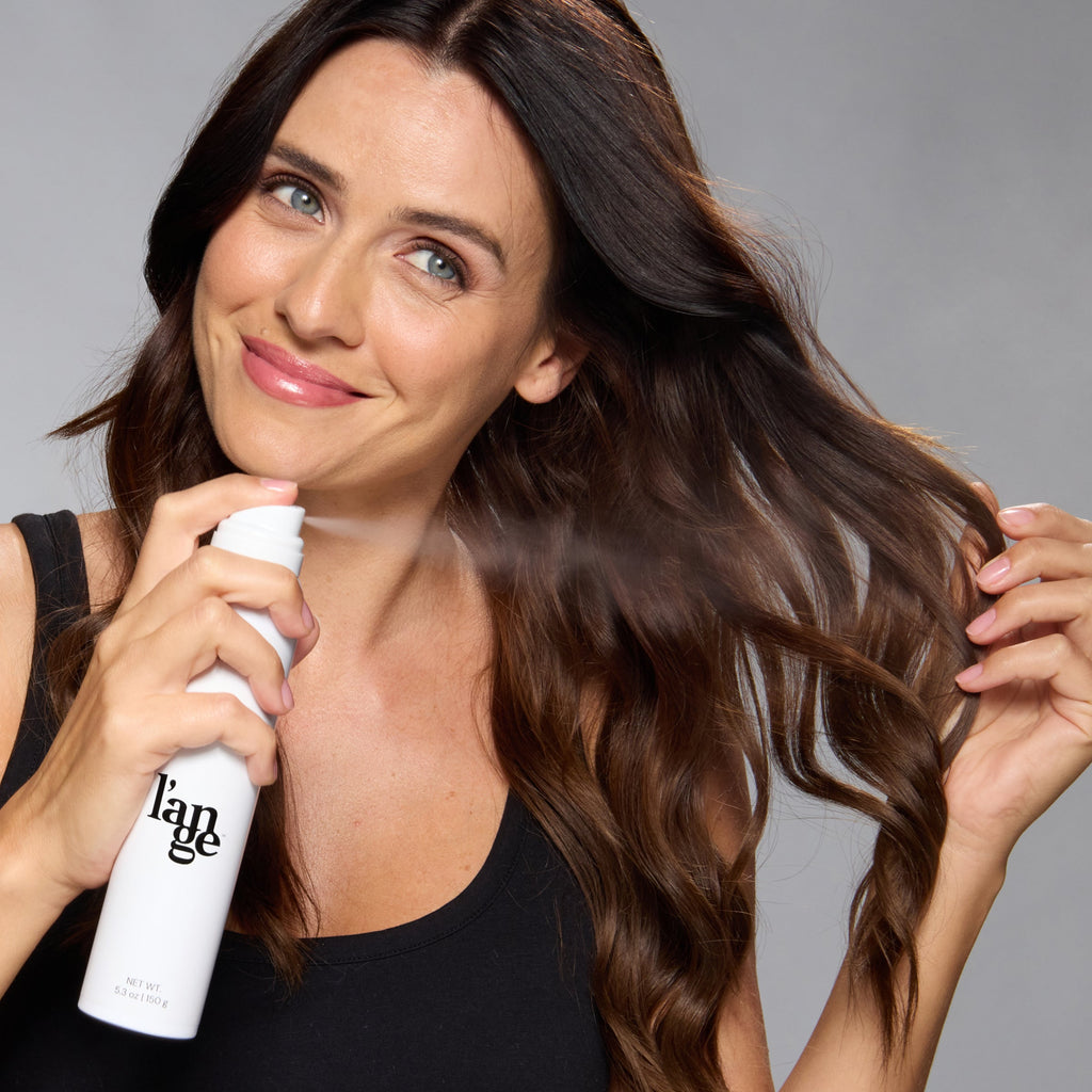 Woman smiling while spraying a white l’ange bottle on her wavy brown hair.