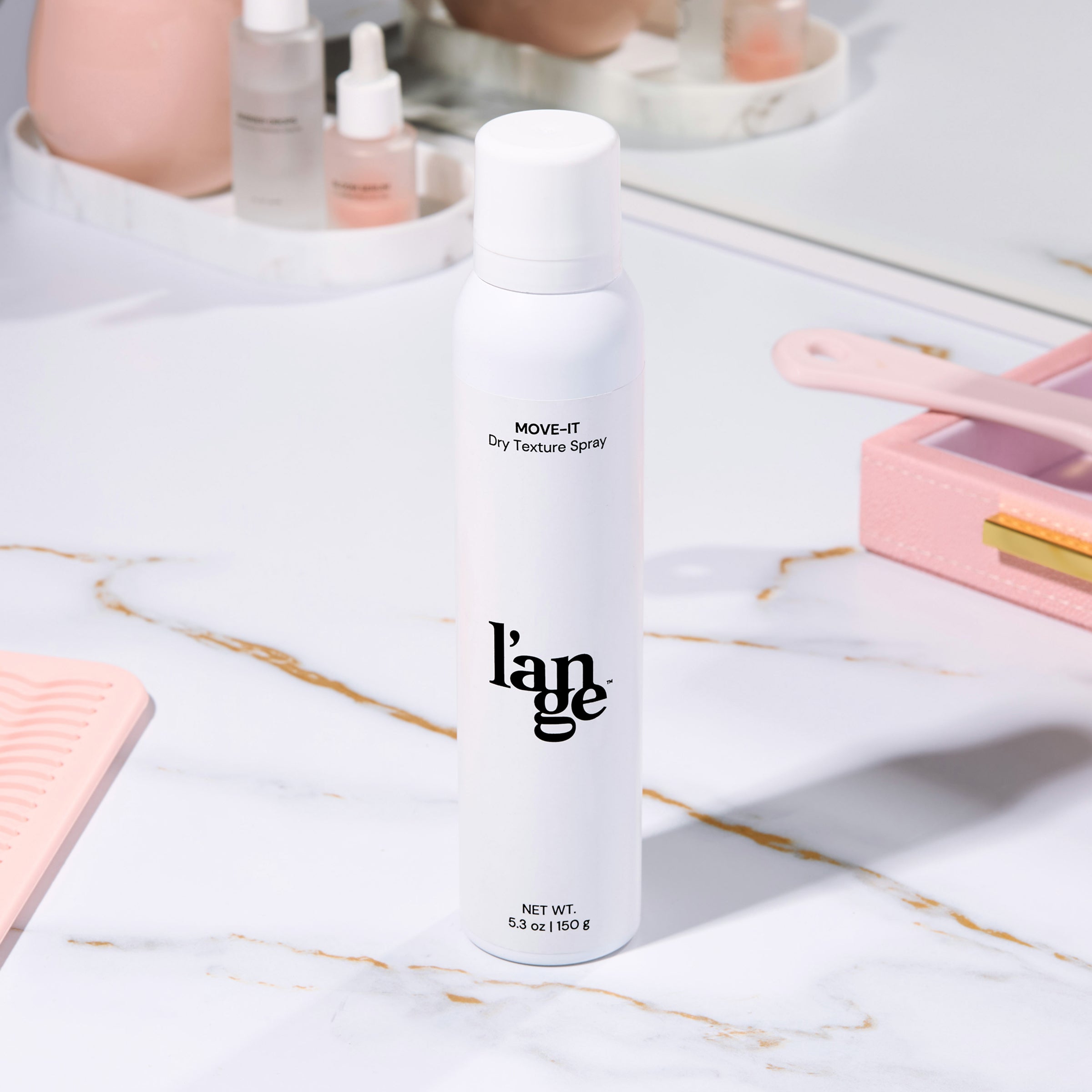 White bottle of l’ange MOVE-IT Dry Texture Spray on a marble vanity surrounded by beauty products.