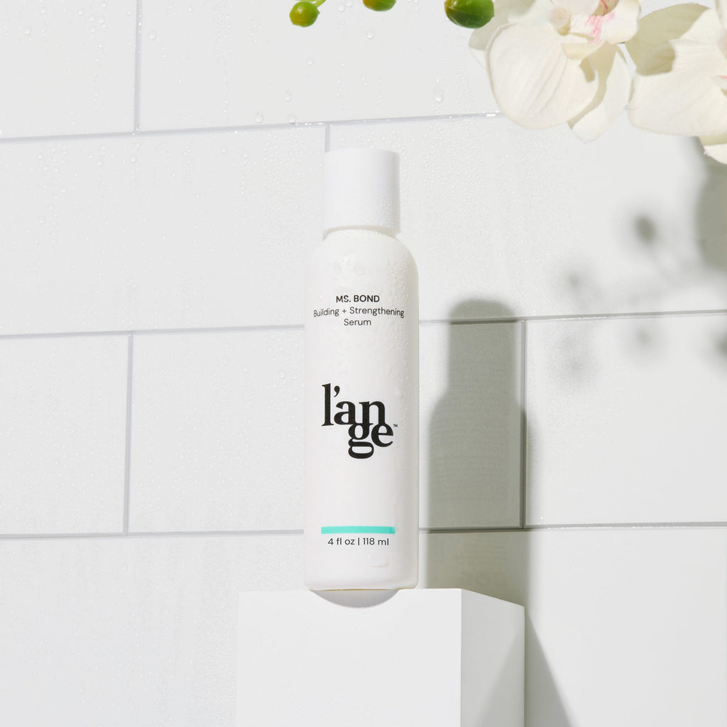 L'ange MS. BOND Building and Strengthening Serum, 4 fl oz (118 ml), displayed in a sleek white bottle against a tiled background with delicate greenery and orchids.