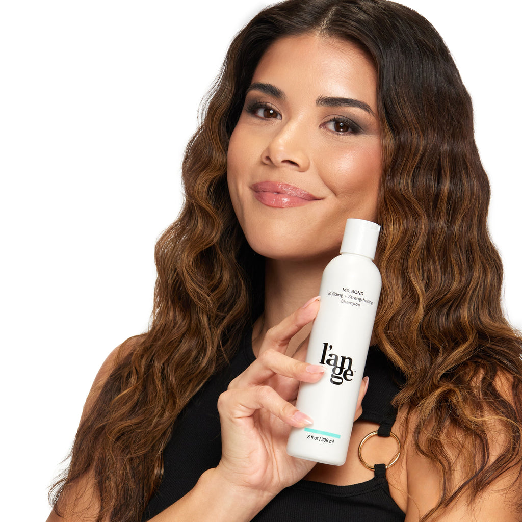 Woman smiling while holding a bottle of l’ange hair strengthening shampoo, showcasing wavy, healthy-looking hair.