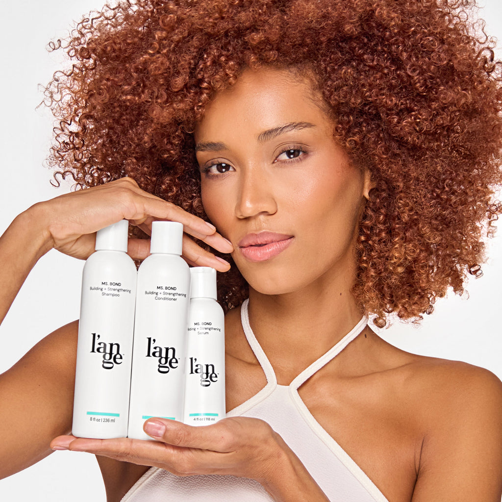 Woman with curly hair holding three hair care products labeled "l’ange" in white bottles: Building + Strengthening Shampoo, Conditioner, and Serum.