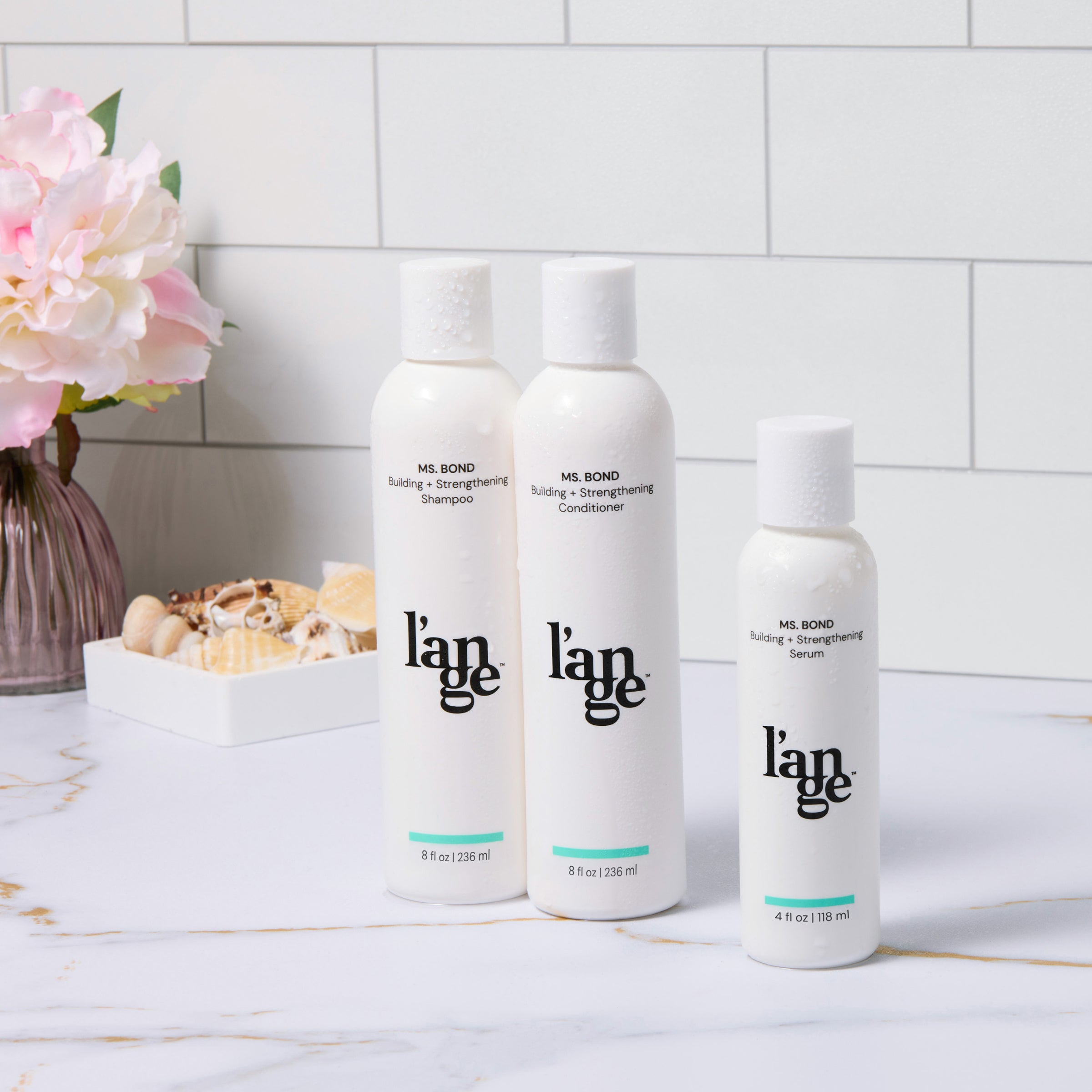 Three bottles of L’ange Ms. Bond hair care products, including shampoo, conditioner, and serum, are elegantly arranged on a marble countertop. A vase with pink and white flowers and a dish of decorative shells are placed in the background, adding a touch of sophistication.