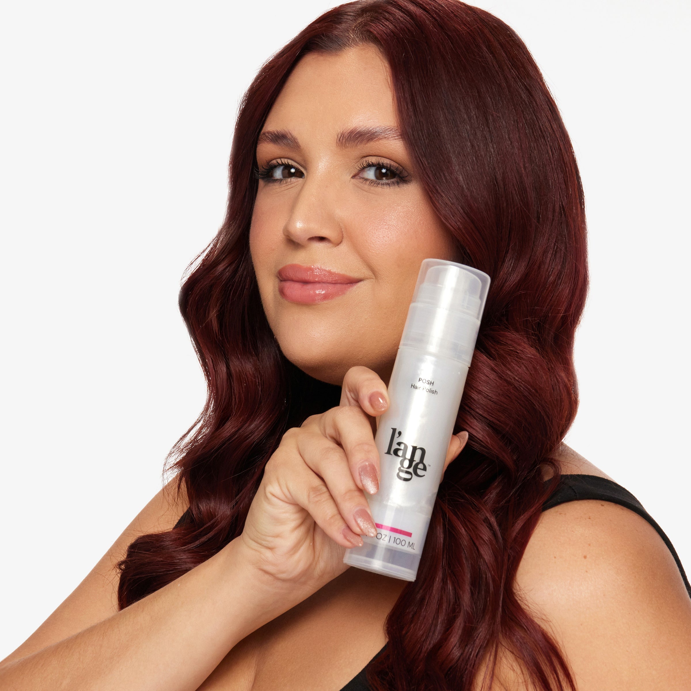 Woman with wavy red hair holding a bottle of L’ange Posh Hair Polish and smiling.