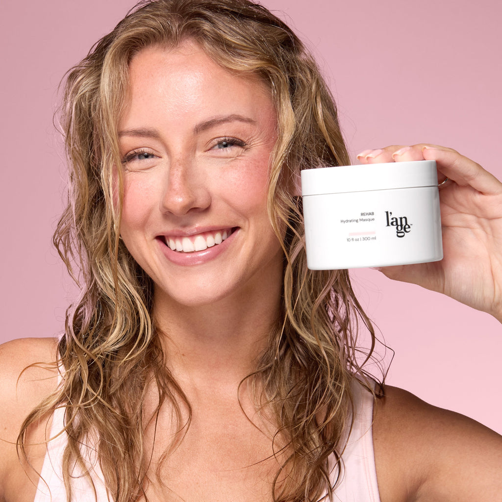 Smiling woman with wavy hair holding a jar of L’ange Rehab Hydrating Masque against a pink background.