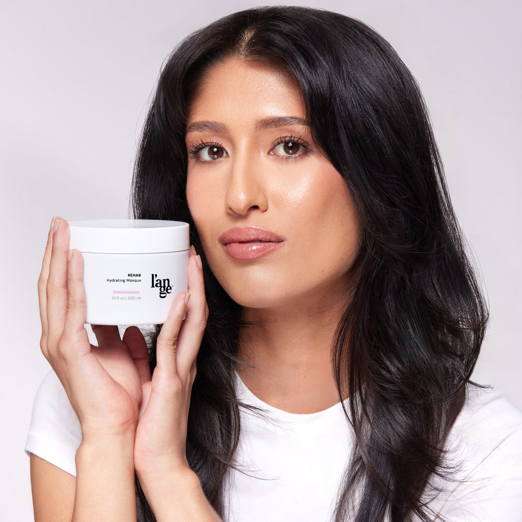 Person with smooth, dark hair holding a white container of L'ange Rehab Hydrating Masque, showcasing the product's design and label.