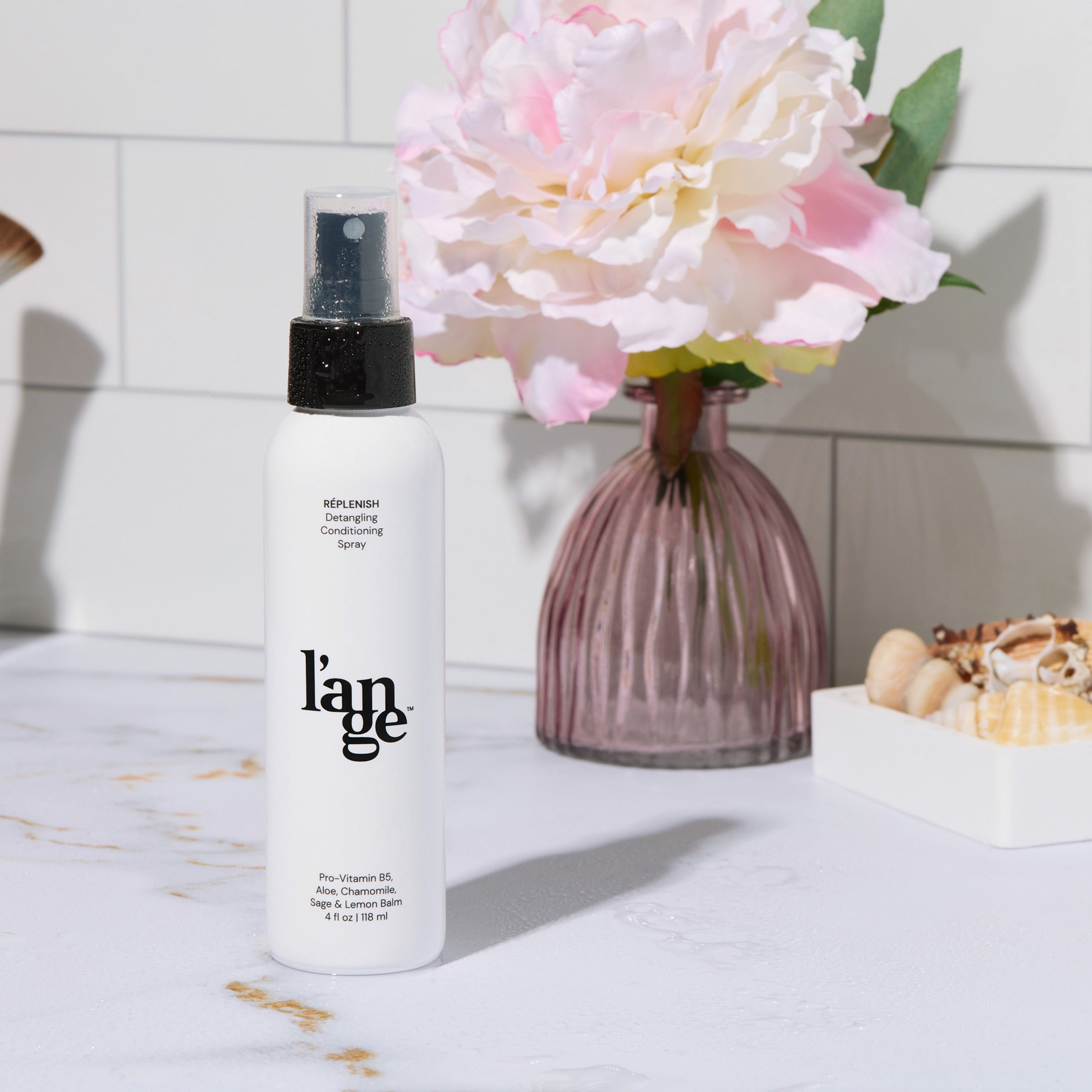 Bottle of L’ange Replenish Detangling Conditioning Spray on a bathroom counter with a pink vase and flower in the background.