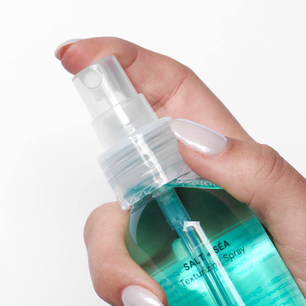 Hand holding a clear spray bottle labeled "Salt + Sea Texturizing Spray" with a blue-green liquid, set against a white background.