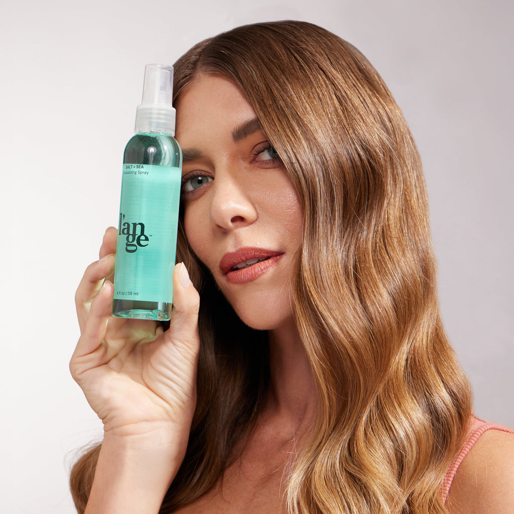 A woman with glossy, honey-blonde waves holds a bottle of Salt + Séa Texturizing Spray next to her face. Her expression is confident, and her hair appears touchably soft and styled in uniform waves.