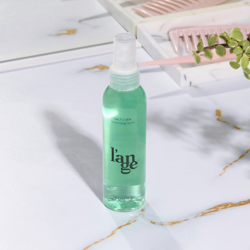 A bottle of L’ange Salt + Séa Texturizing Spray stands upright on a white marble tray in a clean, modern bathroom setting. A white vase and lamp are softly blurred in the background.
