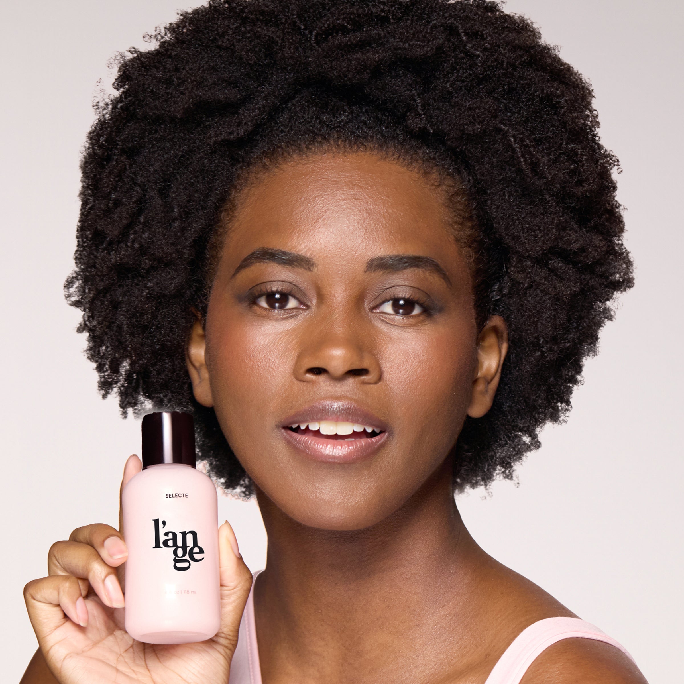 Person with natural afro hairstyle holding a bottle of L'ange hair product, smiling and showcasing the product.