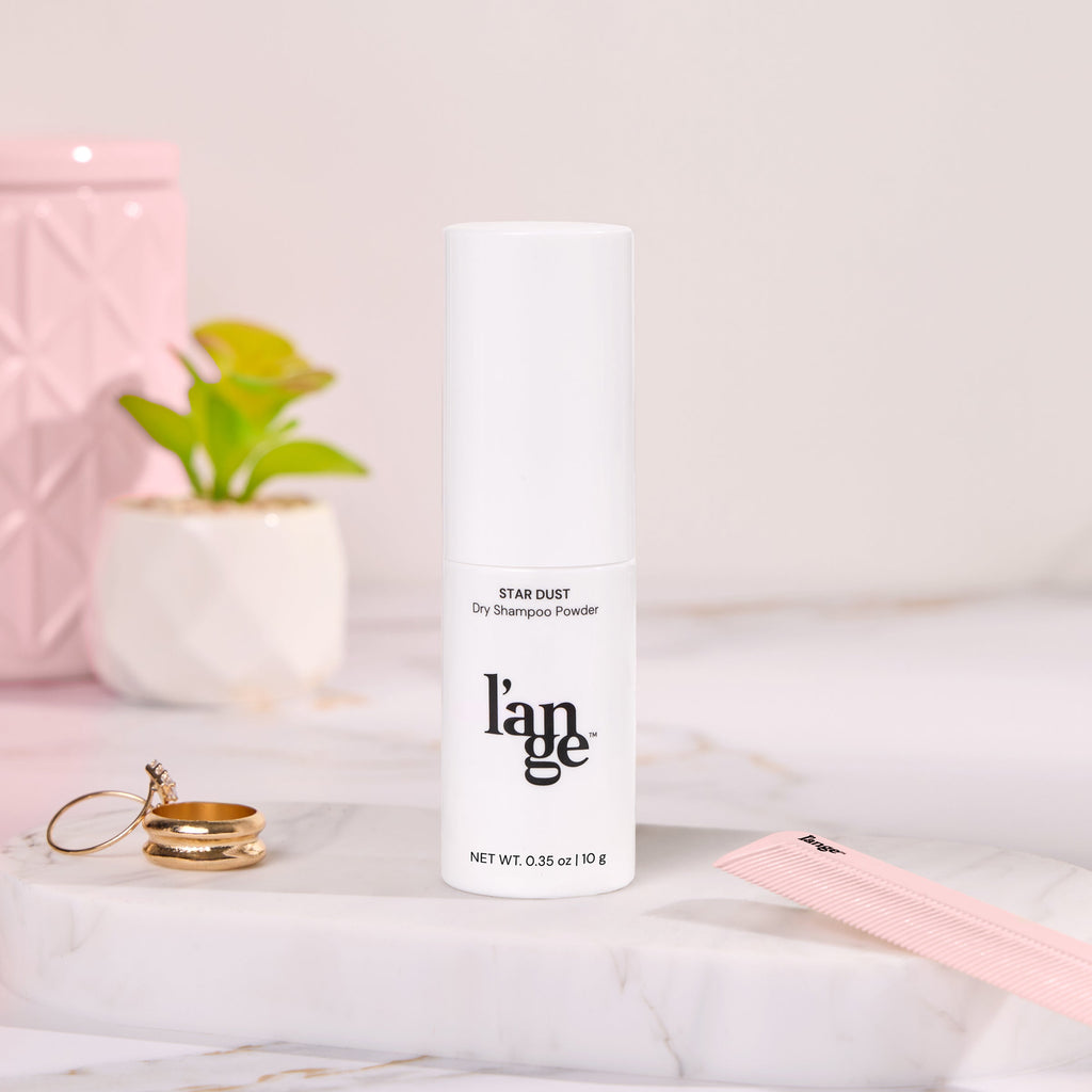 L'ange Star Dust Dry Shampoo Powder in sleek white bottle, surrounded by elegant decor and accessories including a pink comb, gold rings, and small plants on a marble surface.