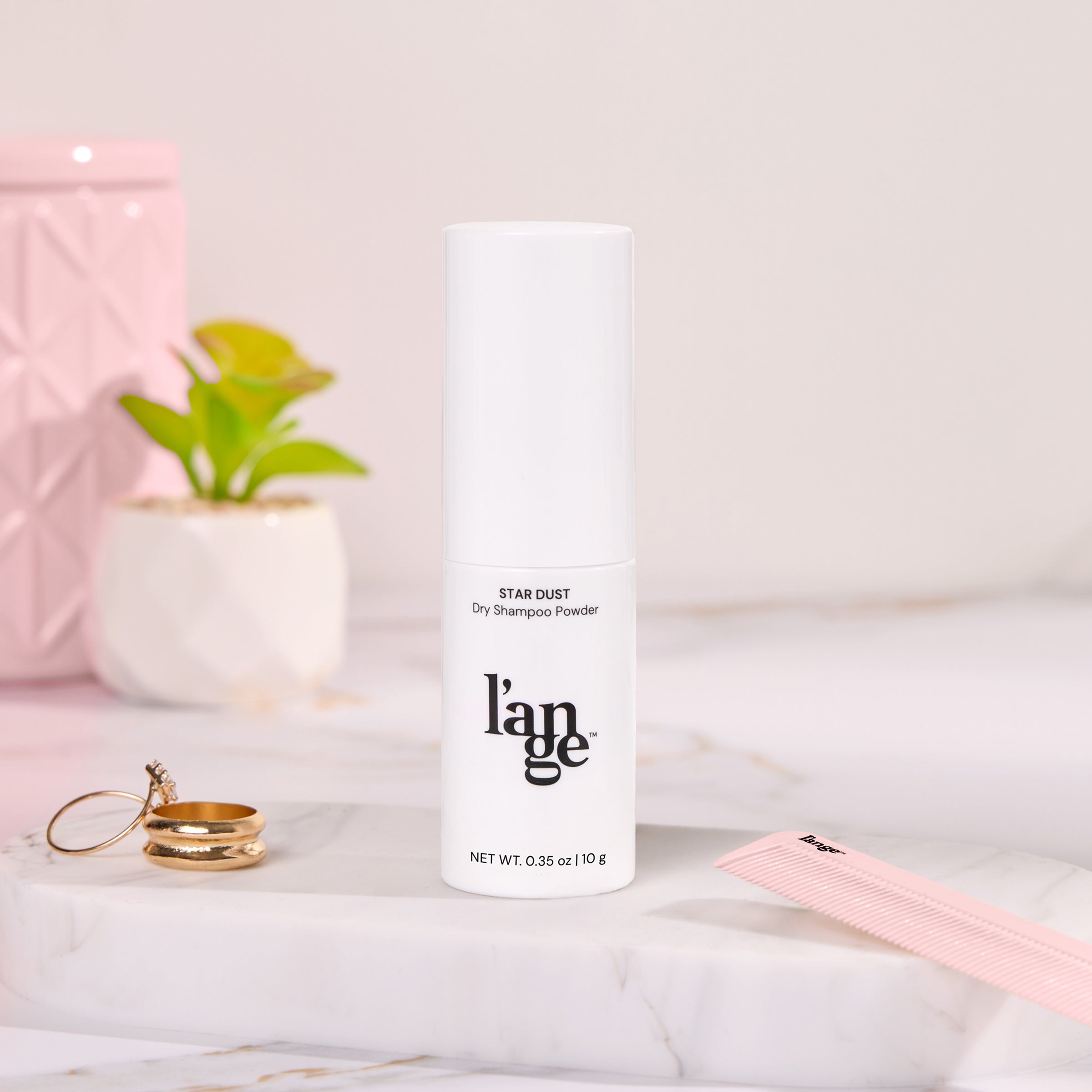 L'ange Star Dust Dry Shampoo Powder in sleek white bottle, surrounded by elegant decor and accessories including a pink comb, gold rings, and small plants on a marble surface.