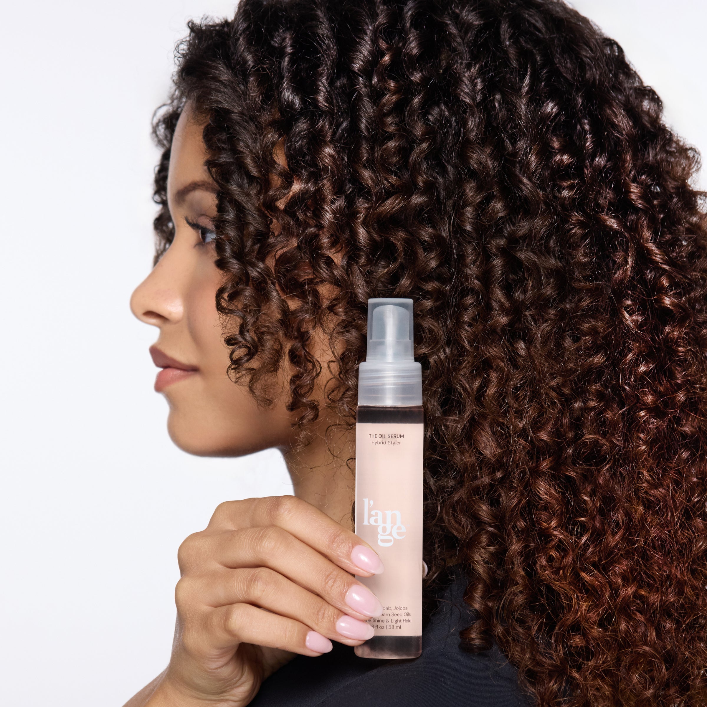 Woman with tight, glossy curls holds a bottle of L’ange The Oil Serum Hybrid Styler next to her face, showcasing defined texture and healthy shine.