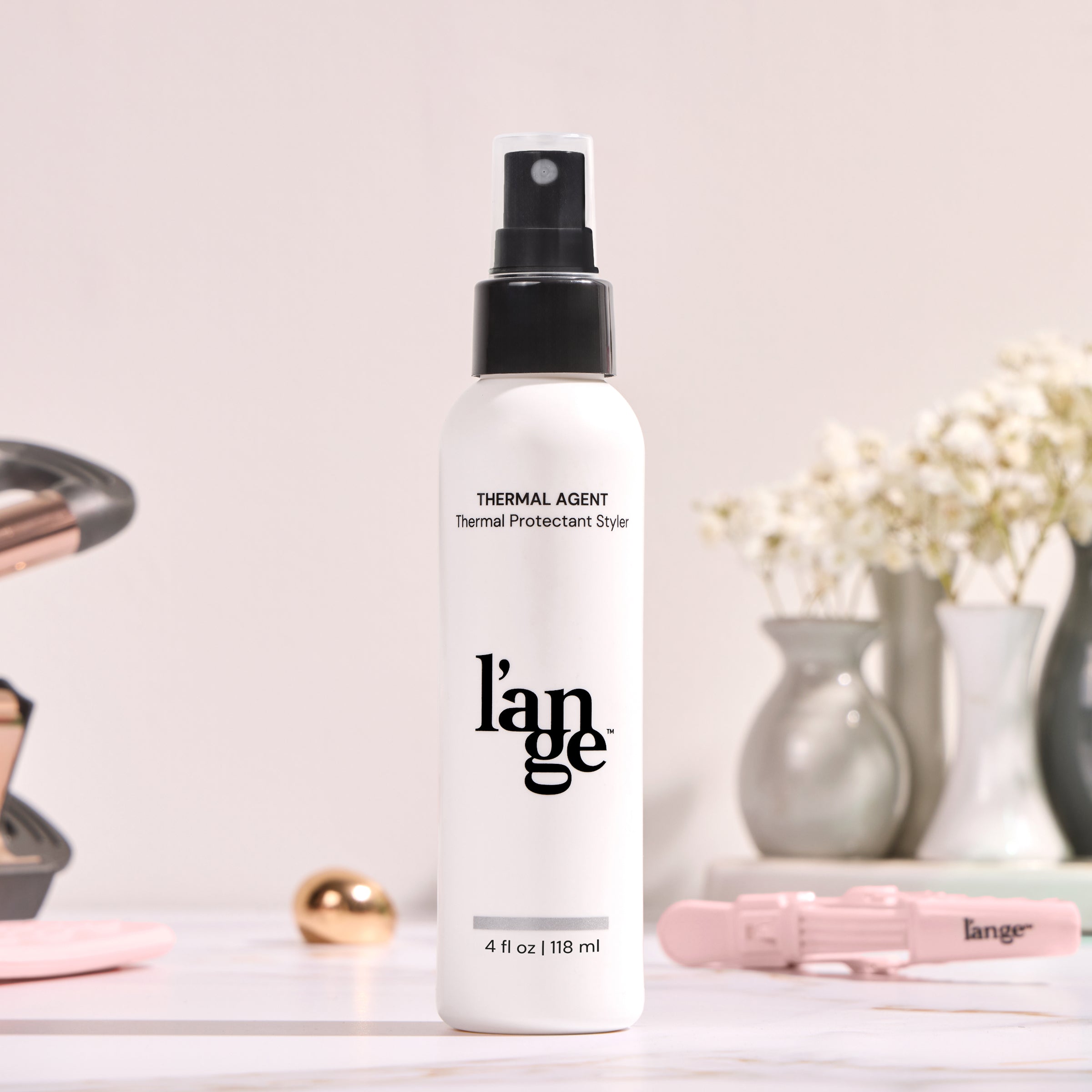 L’ange Thermal Protectant Styler spray, 4 fl oz (118 ml), with a sleek black and white design, positioned on a countertop with blurred styling tools and decorative vases in the background.