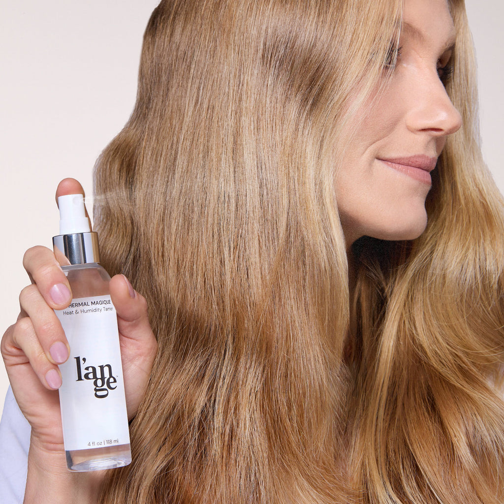 Woman with long, smooth, blonde hair holds a bottle of L’ange Thermal Magique Heat & Humidity Tamer, showcasing sleek, frizz-free strands and a subtle smile.