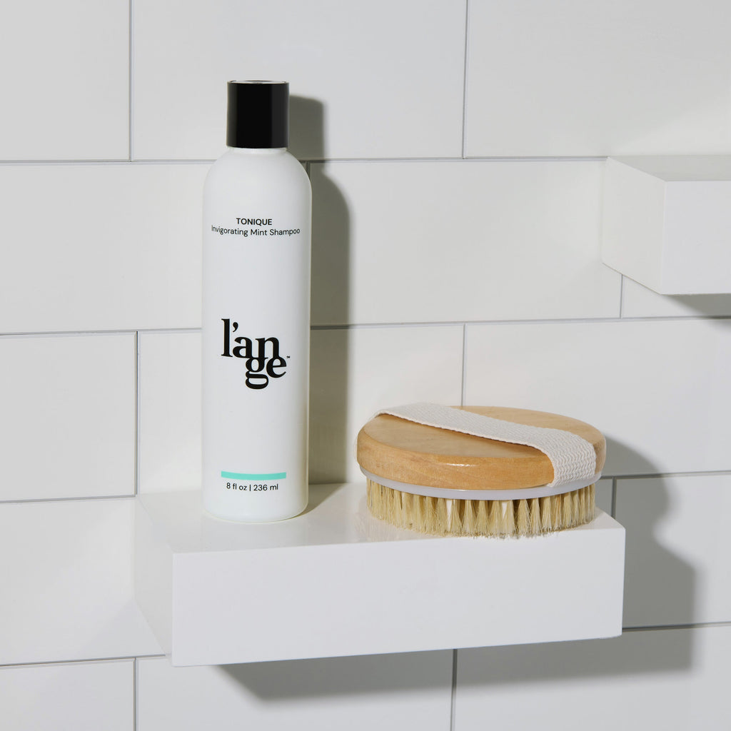 L’ange Tonique shampoo bottle and a dry brush on a shower shelf with white tile background.
